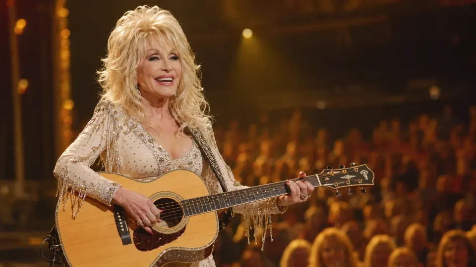 Dolly Parton performing with an acoustic guitar beneath warm golden stage lights.