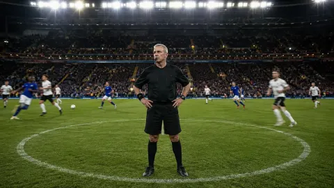 A generic football referee standing near the centre circle while a match continues around him