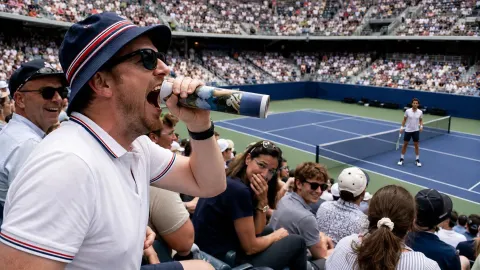Tennis spectator singing through a rolled programme while a player waits on court.