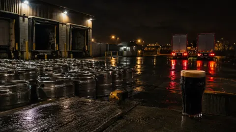 Kegs and two lorries at a dark warehouse loading bay with a pint of stout in the foreground